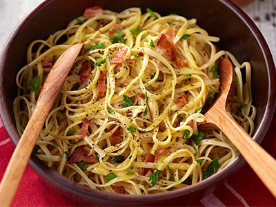 Close-up of spaghetti carbonara with bacon, herbs, and wooden serving utensils.